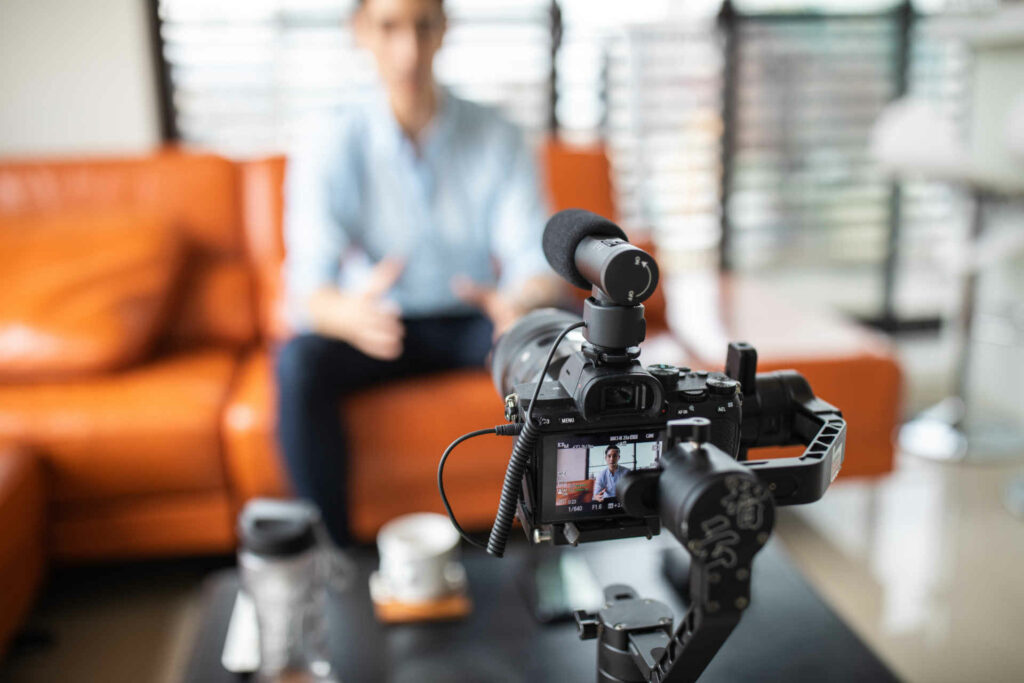 Young handsome man sitting at home on sofa and recording a video with professional camera, looking at camera and talking, focus on foreground